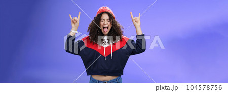 Waist-up shot of rebellious and carefree woman having fun feeling excited and awesome at party close eyes sticking out tongue and showing rock-n-roll signs, wearing beanie and warm clothes 104578756