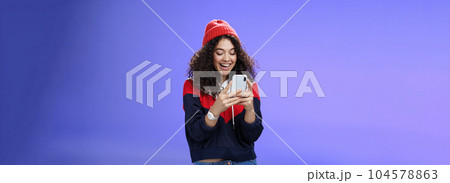 Joyful and sociable stylish caucasian woman with curls in warm beanie having fun laughing and chuckling as using smartphone typing funny message looking at mobile phone screen happily over blue wall Joyful and sociable stylish caucasian woman with curls in warm beanie having fun laughing and chuckling as using smartphone typing funny message looking at mobile phone screen happily over blue wall 104578863