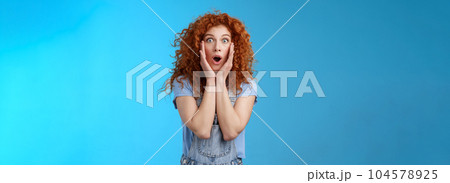Amazed gasping shocked wondered redhead attractive curly girl popping eyes stare camera drop jaw concerned astonished touch cheeks empathy amazement standing blue background Amazed gasping shocked wondered redhead attractive curly girl popping eyes stare camera drop jaw concerned astonished touch cheeks empathy amazement standing blue background 104578925