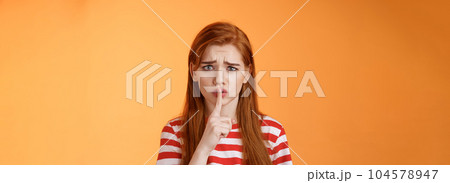 Uneasy worried insecure redhead girl begging keep secret safe, frowning grimace upset, shushing, press index finger lips hush gesture, worry someone spread rumor, orange background 104578947