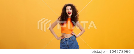 Stylish confident and awesome woman ready lit party standing in trendy cropped top and denim shorts with hands on waist sticking out tongue and smiling joyfully posing over orange background 104578966