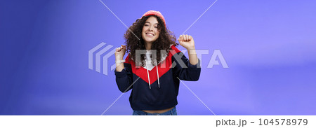 Charming carefree young 20s girl with curly haircut in warm beanie dancing and having fun joyfully over blue wall raising hands relaxed, chill smiling broadly at camera liking perfect chilly weather Charming carefree young 20s girl with curly haircut in warm beanie dancing and having fun joyfully over blue wall raising hands relaxed, chill smiling broadly at camera liking perfect chilly weather 104578979