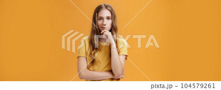 Hmm choices difficult. Portrait of focus serious-looking creative young female with wavy hair smirking holding hand on chin in thoughtful pose, making up decision, thinking over orange background 104579261