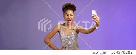 Stylish sassy african american b-day girl taking selfie new elegant silver shiny dress extend arm holding smartphone posing winking display screen amused having fun smiling broadly, blue background Stylish sassy african american b-day girl taking selfie new elegant silver shiny dress extend arm holding smartphone posing winking display screen amused having fun smiling broadly, blue background 104579288