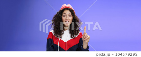 Rule number one do not mess with me babe. Portrait of swag and cool stylish young curly-haired woman in winter beanie showing index finger in prohibition or warning gesture over blue background Rule number one do not mess with me babe. Portrait of swag and cool stylish young curly-haired woman in winter beanie showing index finger in prohibition or warning gesture over blue background 104579295