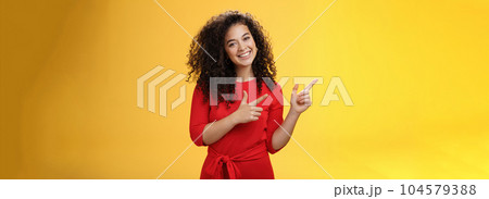 Friendly cute assertive female assistant in elegant red dress with curly hair tilting head flirty and smiling broadly, satisfied as pointing at upper left corner, recommending product over yellow wall Friendly cute assertive female assistant in elegant red dress with curly hair tilting head flirty and smiling broadly, satisfied as pointing at upper left corner, recommending product over yellow wall 104579388