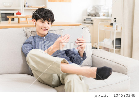 ソファーに座ってイヤフォンをしてタブレットを見て笑っている若い男性 104580349