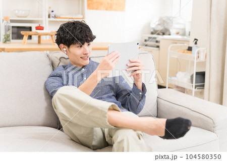 ソファーに座ってイヤフォンをしてタブレットを見て笑っている若い男性 ソファーに座ってイヤフォンをしてタブレットを見て笑っている若い男性 104580350