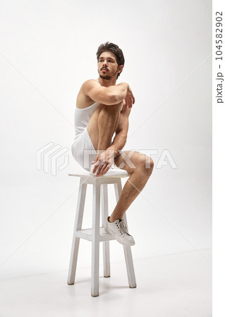 Studio shot of young attractive Caucasian man wearing homewear sitting on high chair over white background. Concept of men's health, beauty of male body, fashion, ad 104582902