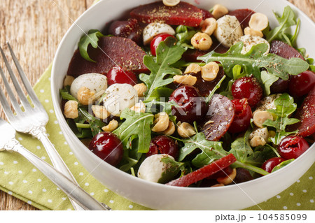 Homemade arugula salad, baked beets, ripe cherries, mozzarella, hazelnuts close-up in a bowl. Horizontal 104585099