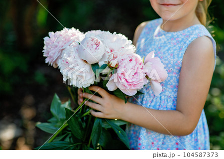 Cute adorable little preschool girl with huge bouquet of blossoming pink peony flowers. Portrait of smiling preschool child in domestic garden on warm spring or summer day. Summertime. 104587373