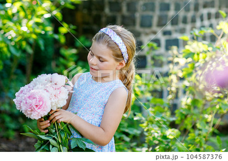 Cute adorable little preschool girl with huge bouquet of blossoming pink peony flowers. Portrait of smiling preschool child in domestic garden on warm spring or summer day. Summertime. Cute adorable little preschool girl with huge bouquet of blossoming pink peony flowers. Portrait of smiling preschool child in domestic garden on warm spring or summer day. Summertime. 104587376