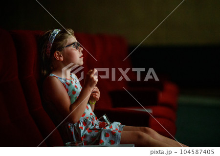 Little preschool girl with glasses watching cartoon movie or film in cinema. Happy excited child. Family activity with children. Little preschool girl with glasses watching cartoon movie or film in cinema. Happy excited child. Family activity with children. 104587405