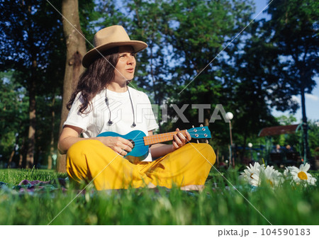 Girl in the park sitting on the grass plays the ukulele. Learning to play a four-string instrument. Relaxation in a public park. Music therapy, psychology, meditation, relaxation, relaxation Girl in the park sitting on the grass plays the ukulele. Learning to play a four-string instrument. Relaxation in a public park. Music therapy, psychology, meditation, relaxation, relaxation 104590183