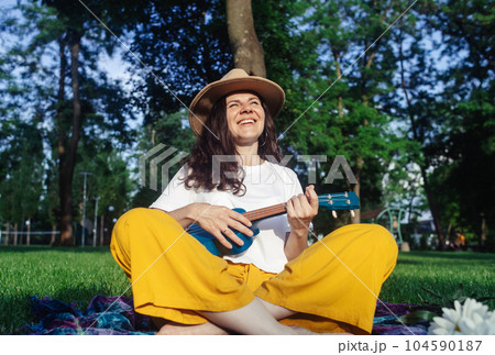 Girl in the park sitting on the grass plays the ukulele. Learning to play a four-string instrument. Relaxation in a public park. Music therapy, psychology, meditation, relaxation, relaxation Girl in the park sitting on the grass plays the ukulele. Learning to play a four-string instrument. Relaxation in a public park. Music therapy, psychology, meditation, relaxation, relaxation 104590187