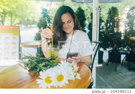 Happy smiling girl sits at a table in a cafe after a romantic date. There is a bouquet of daisies on table. Woman in a brown hat looks at her phone, reads correspondence and remembers happy moments Happy smiling girl sits at a table in a cafe after a romantic date. There is a bouquet of daisies on table. Woman in a brown hat looks at her phone, reads correspondence and remembers happy moments 104590211