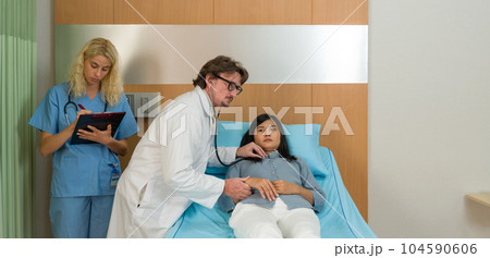 Caucasian doctor holding stethoscope checking heartbeat, examining illness of asian woman patient on the bed in hospital. Young nurse stand next to the bed, writing document on the clipboard. Caucasian doctor holding stethoscope checking heartbeat, examining illness of asian woman patient on the bed in hospital. Young nurse stand next to the bed, writing document on the clipboard. 104590606