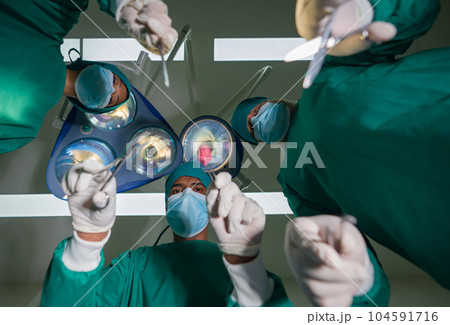 Emergency surgical care concept. Group of surgeons and nurse in surgical green gown uniform performing surgical operation in operating room. Bottom view. Emergency surgical care concept. Group of surgeons and nurse in surgical green gown uniform performing surgical operation in operating room. Bottom view. 104591716