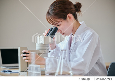 顕微鏡のレンズを覗いて観察する白衣の若い女性 顕微鏡のレンズを覗いて観察する白衣の若い女性 104602794