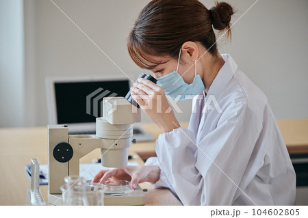 顕微鏡のレンズを覗いて観察する白衣の若い女性 104602805