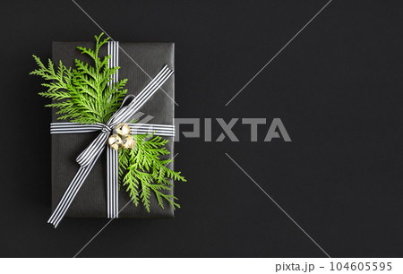 Black gift box with sprig of thuja and bells on black background. Space for your text. Black gift box with sprig of thuja and bells on black background. Space for your text. 104605595