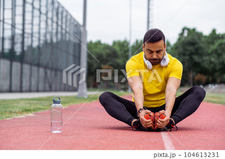 Active sportsman in earphones stretches with the butterfly exercise on the track at a large stadium in a green city park's urban sports arena.  104613231