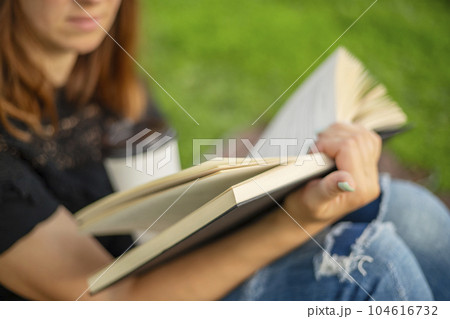 A woman sits near a tree in the park and holds a book and a cup with a hot drink 104616732