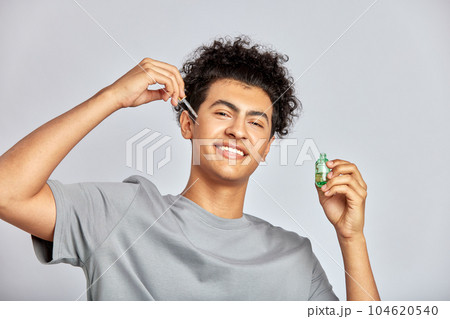 Young guy with black curly hair moisturizing his skin with a facial oil in an anti-aging skincare routine. Handsome swarthy man pampering his skin to a achieve a more healthy and glowing appearance. Young guy with black curly hair moisturizing his skin with a facial oil in an anti-aging skincare routine. Handsome swarthy man pampering his skin to a achieve a more healthy and glowing appearance. 104620540