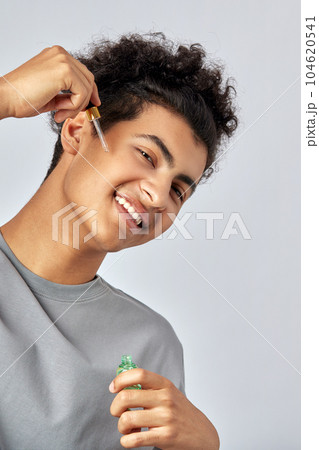 Young guy with black curly hair moisturizing his skin with a facial oil in an anti-aging skincare routine. Handsome swarthy man pampering his skin to a achieve a more healthy and glowing appearance. Young guy with black curly hair moisturizing his skin with a facial oil in an anti-aging skincare routine. Handsome swarthy man pampering his skin to a achieve a more healthy and glowing appearance. 104620541