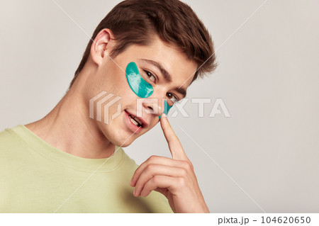 A millennial guy with brown hair practicing skincare with under eye hydrogel patches in a studio. Young smiling caucasian man applying cosmetic patches on his face. Men's self-care. 104620650