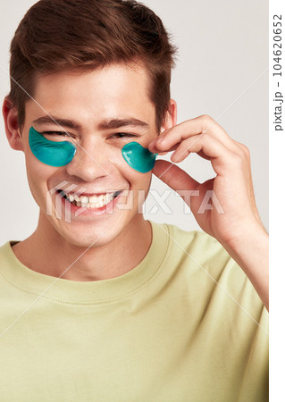 A millennial guy with brown hair practicing skincare with under eye hydrogel patches in a studio. Young smiling caucasian man applying cosmetic patches on his face. Men's self-care. 104620652