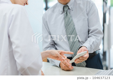 薬局で高齢者・中高年男性にスマホアプリを説明する薬剤師・調剤師の女性（服薬指導） 104637123