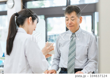 薬局で高齢者・中高年男性にスマホアプリを説明する薬剤師・調剤師の女性（服薬指導） 104637124