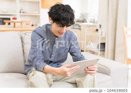 ソファーに座って,かがんでイヤフォンをしてタブレットを見て笑っている若い男性 104639005