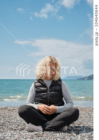 Adult tourist european woman blonde sitting alone on a pebbly beach with blue ocean and sky in the background, vertical shot. People and summer holidays vacation lifestyle concept 104640644