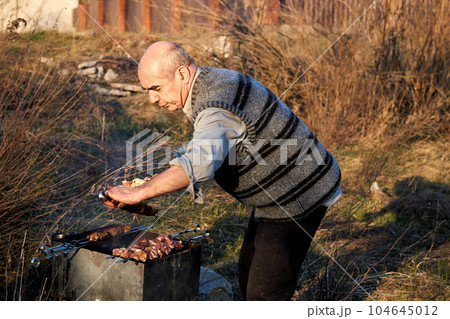 Elderly cheerful man pensioner cooking meat on the grill outdoors in nature 104645012