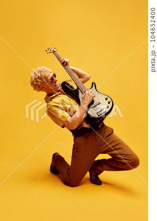 Artistic, expressive man with blonde curly hair, in sunglasses and stylish clothes emotionally playing guitar against yellow studio background Artistic, expressive man with blonde curly hair, in sunglasses and stylish clothes emotionally playing guitar against yellow studio background 104652400