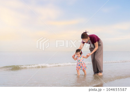 Asian mother holding hand daughter for support and walking on beach, Baby girl learning to walk, Happy family activity concept. 104655587