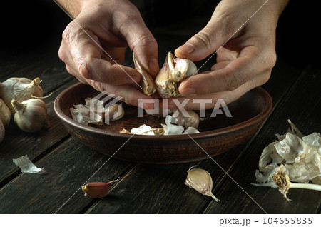 Close-up of a cook hands peeling garlic in the kitchen. The concept of cooking vegetarian food on a dark background 104655835
