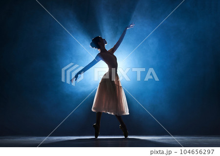 Silhouette of tender, graceful young woman, ballerina performing, dancing over dark blue background with spotlight 104656297