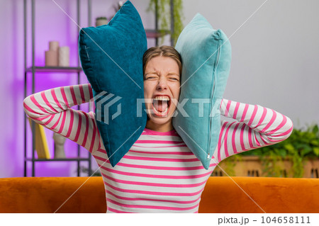 Repair work at neighbours concept, irritated woman at home cover ears with pillows annoyed by noise Repair work at neighbours concept, irritated woman at home cover ears with pillows annoyed by noise 104658111
