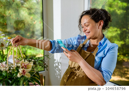 Smiling pregnant woman florist photographing a bouquet of orchid flowers in a wicker basket, sharing it on social media Smiling pregnant woman florist photographing a bouquet of orchid flowers in a wicker basket, sharing it on social media 104658796