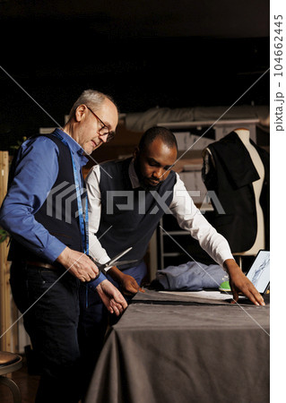 Professional suitmaker and apprentice cutting textile material with scissors in tailoring studio, designing upcoming attire collection. Fashion industry craftmen manufacturing suit in atelier 104662445