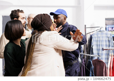 Male security guard pushing mad diverse people shoppers back while controlling clothing store entrance before Black Friday sales. Anxious and angry customers waiting in line for Cyber Monday discounts 104662475