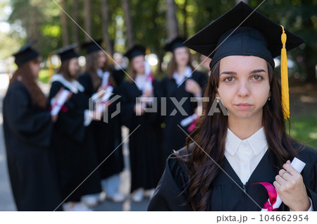 Group of happy students in graduation gowns outdoors. A young girl is holding a diploma. 104662954