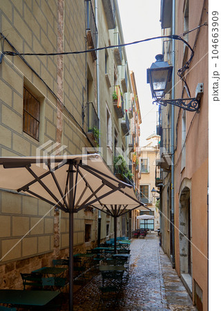 Narrow street Girona. Narrow medieval street in the Jewish quarter in Girona, Catalonia, Spain. An old atmospheric city with narrow and cramped quiet streets. Сolourful narrow alleys in the old town 104663909