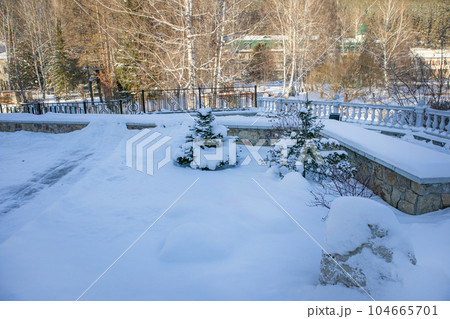 A snow-covered park with a stone balustrade and fence is visible, the house is visible through the trees in the background, in a charming winter landscape A snow-covered park with a stone balustrade and fence is visible, the house is visible through the trees in the background, in a charming winter landscape 104665701