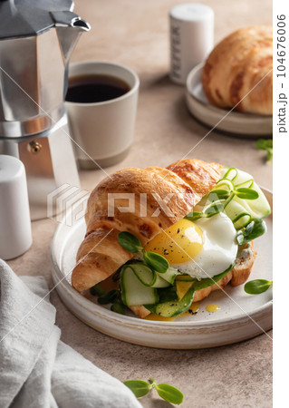 Delisious breakfast with croissant sandwich with fried egg, cheese, cucumber and micro greens and coffee over beige background. Healthy delicious breakfast 104676006