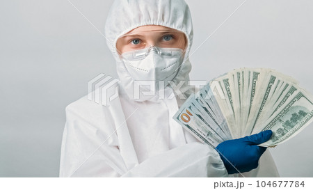 Healthcare salary. Doctor earnings. Female medical professional woman in white ppe blue gloves with dollar money fan isolated on grey background. 104677784