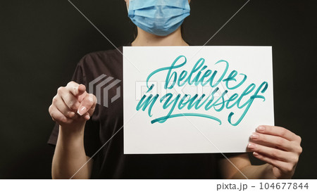 Believe in yourself. Life motivation. Unrecognizable woman in face mask pointing finger showing supportive slogan placard on black copy space. Believe in yourself. Life motivation. Unrecognizable woman in face mask pointing finger showing supportive slogan placard on black copy space. 104677844
